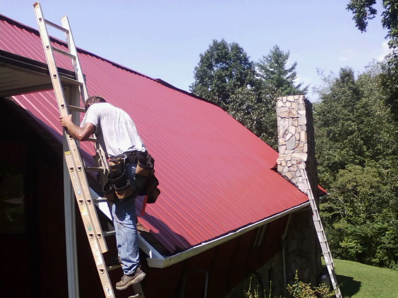 Roofer on a ladder beside a red metal roof and stone chimney — Soffit & Fascia Repair by Roofing Brothers in Meldrim